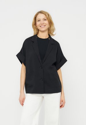 Usměvavá žena s blond vlasy, která nosí volný černý krátký sakó přes černý top, doplněné bílými kalhotami s širokými nohavicemi.