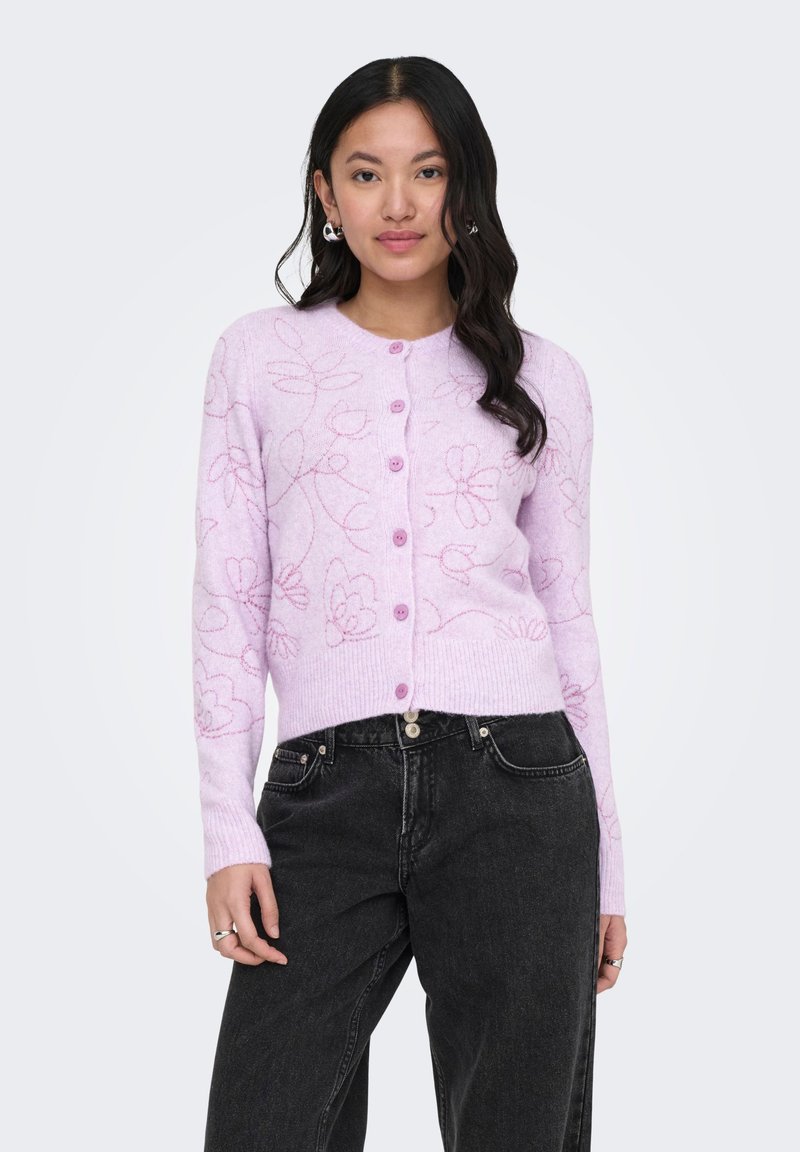 Jeune femme aux longs cheveux foncés portant un cardigan boutonné à motif floral violet clair et un jean noir taille haute, debout dans une pose détendue.