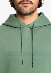 Zelená mikina s kapucňou vyrobená z mäkkej látky, s predným logom "DC Shoes" v tmavšej zelenej farbe. Obsahuje nastaviteľné šnúrky s kovovými koncami.