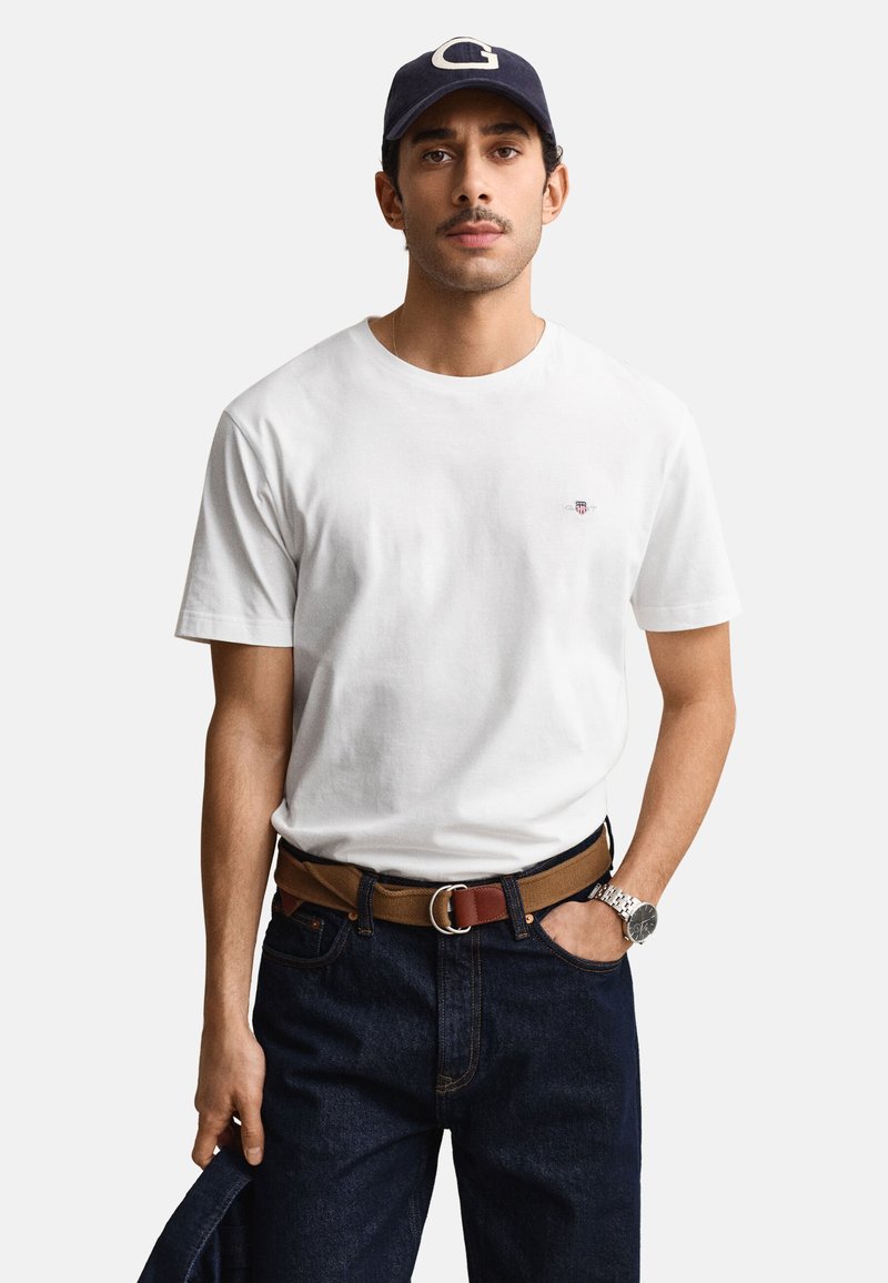 Uomo che indossa una maglietta bianca, jeans scuri, una cintura marrone, un cappellino blu navy e un orologio da polso argentato, che tiene una giacca scura in una mano.