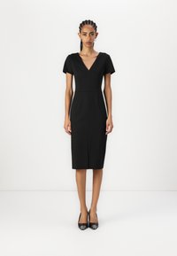 Black fitted dress with short sleeves, V-neckline, and knee-length hem. Made from smooth fabric with subtle stitching detail. Black heels.