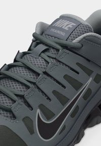 Κοντινό πλάνο γκρι αθλητικού παπουτσιού Nike με επάνω μέρος από δίχτυ, μαύρο λογότυπο Swoosh και σκούρες γκρι κορδόνια περασμένα μέσα από συνθετικούς θηλιές.