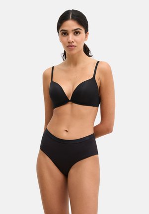 Frau mit dunklen, zurückgebundenen Haaren, die einen schwarzen BH und hoch taillierte schwarze Unterwäsche vor einem schlichten hellen Hintergrund trägt.