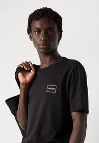 Fiatal férfi, rövid rasztahajjal, fekete HUGO pólót visel, és egy fekete dzsekit tart a válla fölött, egy sima világos háttér előtt.