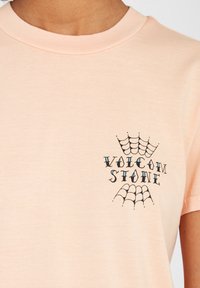 Perzikkleurig katoenen T-shirt met ronde hals; bevat een zwart spinnenwebgraphic en de tekst "VOLCOM STONE" in gestileerd lettertype.