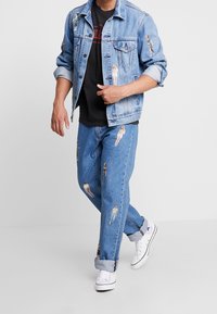 Chaqueta de mezclilla de color azul claro con parches de figuras, combinada con unos vaqueros de mezclilla azul claro a juego que presentan parches similares. Camiseta negra y zapatillas blancas.