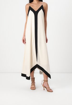 Kobieta ubrana w kremową sukienkę midi bez rękawów z czarnym geometrycznym wykończeniem oraz jasnobrązowe sandały na obcasie na cienkich paskach na jednolitym białym tle.