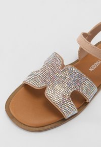 Sandale plate avec une tige en daim beige, dotée d'une grande sangle décorative recouverte de petites strass irisés. Semelle marron.