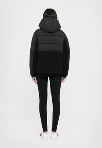 Zwarte pufferjack met een capuchon, met een fluffy zwarte ondersectie. Draagt het met zwarte leggings en witte sneakers, gezien vanuit de achterkant.