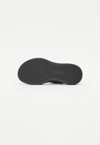 Semelle en caoutchouc noir d'une chaussure avec un motif de bande de roulement texturé et le logo de la marque embossé au centre, photographiée sur un fond blanc.