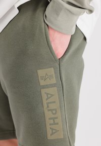 Shorts vert olive en tissu doux en mélange de coton. Présente un patch avec le texte "ALPHA", une poche avant et une taille élastique.