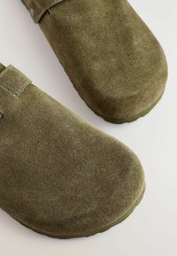Olive green suede slip-on shoes with rounded toes, stitched accents, and flat rubber soles. Smooth texture with no visible patterns.