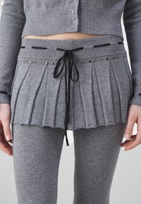 Cárdigan gris corto de punto acanalado y minifalda plisada a juego con cordón negro y tachuelas plateadas, sobre leggings.