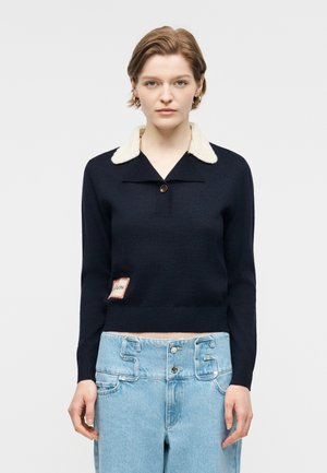 Νεαρή γυναίκα με κοντά μαλλιά φοράει ένα μπλε πουλόβερ με λευκό γιακά και ανοιχτόχρωμο τζιν με μοναδική ζώνη μέσης με διπλό κουμπί.