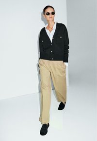 Cardigan nero a maglia con due tasche, chiuso con bottoni. Camicia bianca sotto, abbinata a pantaloni beige a gamba larga e scarpe nere.