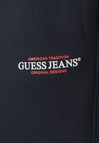 Ciemnogranatowa tkanina z logo "GUESS JEANS" w bieli z czerwonymi akcentami. Zawiera tekst "AMERICAN TRADITION" oraz "ORIGINAL DESIGNS."