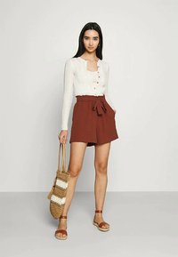 Nainen, jolla on päällään valkoinen napillinen neuletakki ja ruosteenväriset sidotut shortsit, pitää kädessään raidallista kudottua laukkua seisten yksinkertaista taustaa vasten.