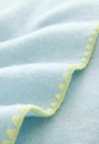 Svetlomodrá flísová tkanina s vlnitým okrajom zo žltej prešívanej niťe. Textúra je mäkká a hladká, s jemnými farebnými odtieňmi.