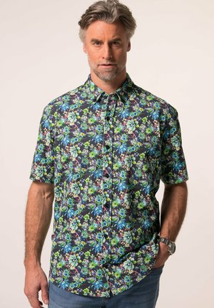 Uomo con capelli grigi che indossa una camicia a maniche corte con bottoni e motivo floreale colorato su uno sfondo uniforme.