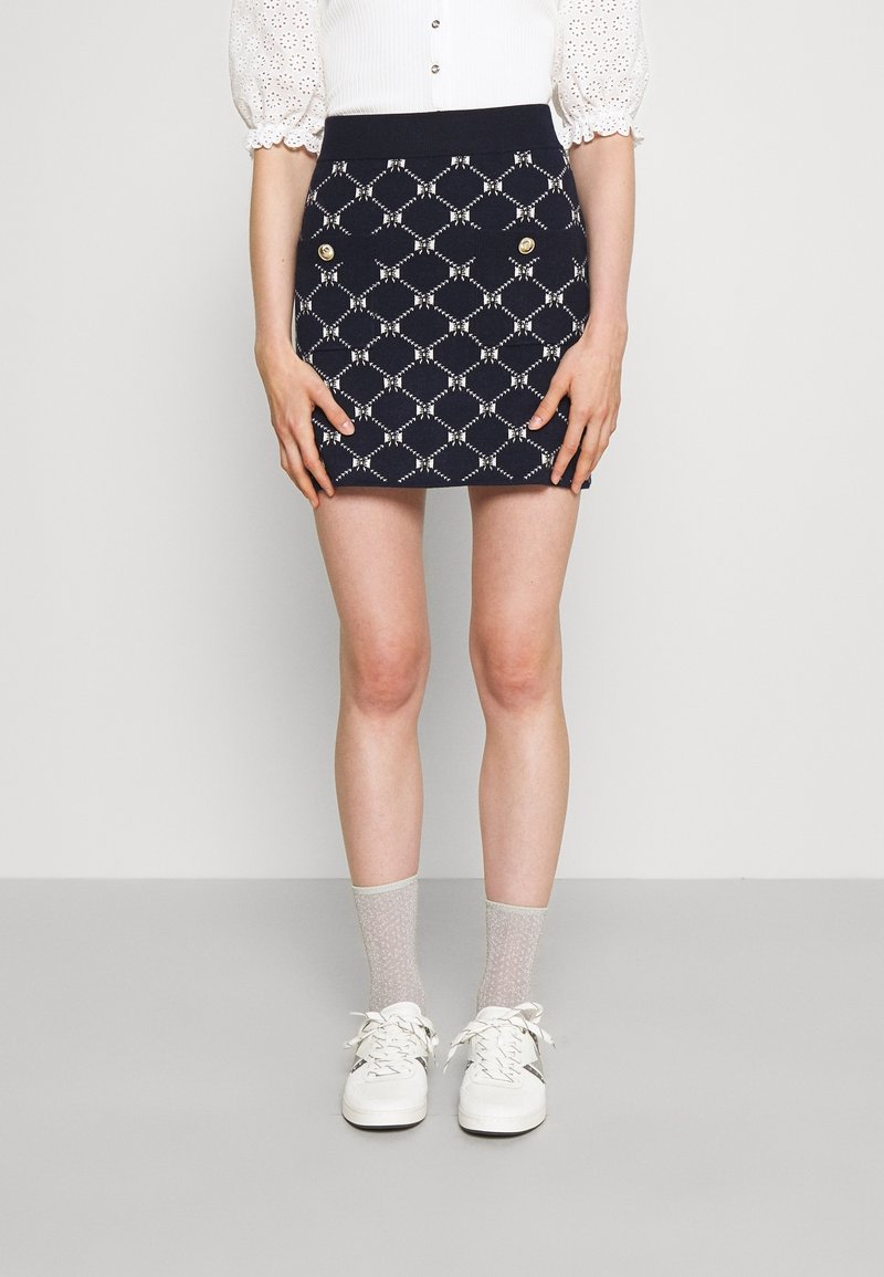 Jupe mini marine avec un motif de diamants blancs et des boutons dorés à l'avant, fabriquée à partir d'un tissu texturé. Portée avec des chaussettes grises à motifs et des baskets.