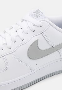 Hvide lædersneakers med grå Nike swoosh-logo, hvide snørebånd og struktureret sål. Har syninger og et mærket tunge-label.