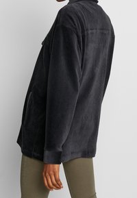 Chaqueta de pana negra con cuello, bolsillos laterales, corte relajado y textura acanalada, combinada con pantalones verde olivo.