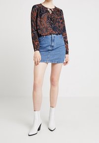 Blusa negra con patrón de paisley naranja y escote de ojal, combinada con una falda corta de mezclilla azul y botas de tobillo blancas con tacón.