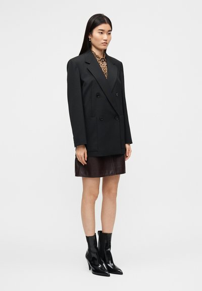  Μαύρο μπλέιζερ διπλής σειράς με μεγάλες τσέπες, φορεμένο πάνω από καφέ μίνι φούστα από δέρμα. Το μοντέλο φοράει μαύρες μυτερές μποτάκια αστραγάλου.