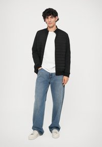 Jacket preta acolchoada com fecho de correr e gola alta, usada sobre uma t-shirt branca. Jeans de corte largo em azul claro e ténis brancos completam o visual.
