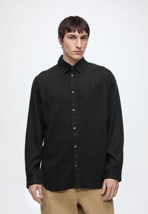 Homme portant une chemise noire à manches longues avec un petit logo sur la poitrine et un pantalon beige, sur un fond blanc uni.