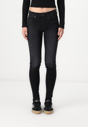 Personne portant un jean skinny noir, un crop top noir à manches longues et des baskets Adidas noires avec des bandes blanches et des semelles en gomme.