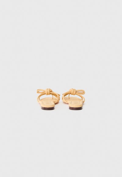 Des sandales beiges avec des brides nouées et une semelle plate. Le matériau supérieur semble texturé, avec un design simple et un matériel minimal.