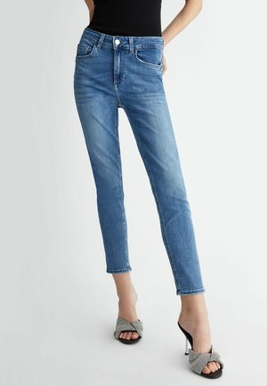 Frau trägt mittel-hohe blaue Skinny-Jeans und schwarze Sandalen mit hohen Absätzen und geknoteten Riemen, stehend vor einem schlichten Hintergrund.