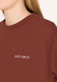 Mörkröd t-shirt med rund halsringning, prydd med vit broderad text "LES'ARCS" på framsidan. Tillverkad av ett mjukt och slätt tyg.