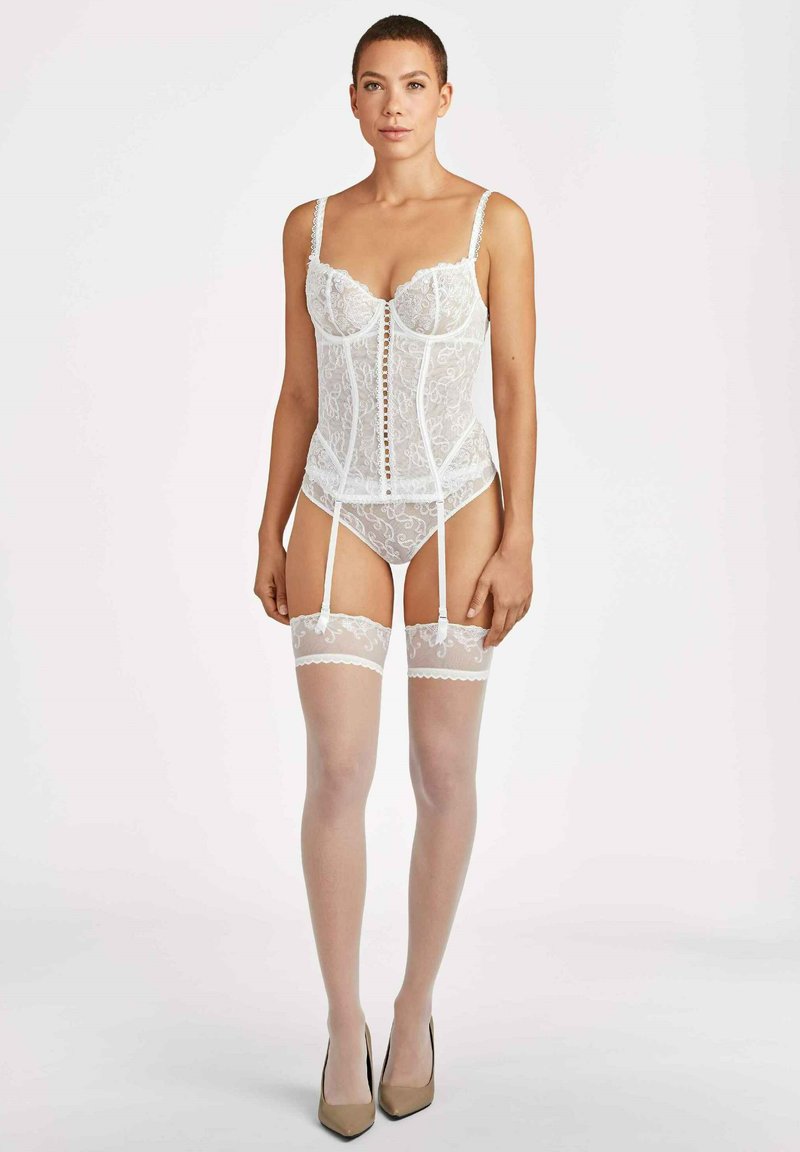 Corset en dentelle blanche avec un motif floral, bonnets à armature, bretelles réglables et fermeture à boutons sur le devant, accompagné de bas transparents montants jusqu'à la cuisse.