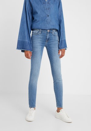 Person trägt hellblaue Skinny-Jeans, Denim-Hemd mit weiten Ärmeln und weiße Sneaker, steht vor einem schlichten hellen Hintergrund.