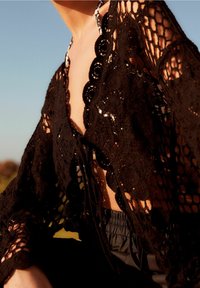 Osoba w czarnym szydełkowym pokrowcu na kostium kąpielowy, siedząca na zewnątrz w ciepłym świetle słonecznym i pod czystym niebem.