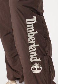 Brown nylon pants featuring a vertical "Timberland" logo and tree emblem in cream. Smooth texture with side seams and elastic cuffs.