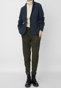 Blazer azul marino de tela texturizada, con dos bolsillos frontales, usado sobre un cuello de tortuga crema. Pantalones verde oliva ajustados con cremalleras, combinados con botas negras.