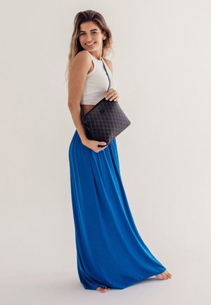 Schwarz gemusterte Umhängetasche, gehalten von einer Person, die ein weißes Crop-Top und einen fließenden blauen Rock trägt, vor einem hellen Hintergrund.