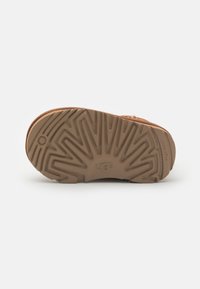 Beige Gummi-Schuhsohle mit strukturiertem Muster, das das eingestempelte "UGG"-Logo in der Mitte und ein kreisförmiges Profilierungsdetail aufweist.