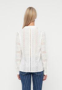 White blouse with long sleeves, featuring eyelet cut-out patterns and a straight hem. Cotton material with a collared neckline.