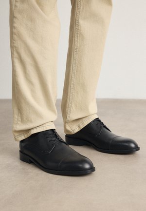 Black leather dress shoes with a rounded toe, textured surface, and laces, paired with light beige pants, standing on a light gray floor.