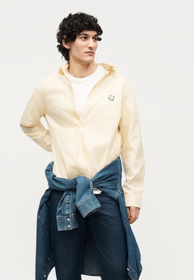 Νέος άνδρας με σγουρά μαλλιά που φορά ανοιχτόκίτρινο πουκάμισο πάνω από λευκό μπλουζάκι, μπλε τζιν και τζιν μπουφάν δεμένο στη μέση του.