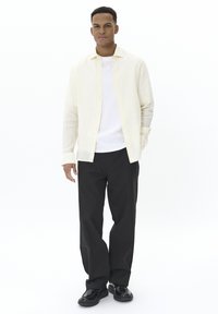 Cămașă cu dungi galbene deschise și albe, peste un tricou alb, pantaloni negri și pantofi negri, cu mâinile în buzunare, pe un fundal neutru.