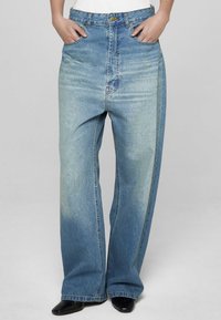 Široke modre jeans hlače z lahkim pranjem, opremljene s sprednjimi žepi, zapiranjem na gumbe in rahlo bledenim dizajnom blizu roba.