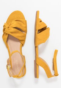 Korkeakorkoiset keltaiset nahkasaappaat, joissa on solmittu etuosa, nilkkahihna ja litteä pohja. Sileä pinta vähäisellä varustuksella.