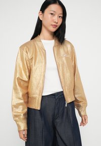 Jacket bombă metalic aurie cu manșete și guler ribbate, cu fermoar frontal și logo brodat pe piept. Purtați peste un top alb.