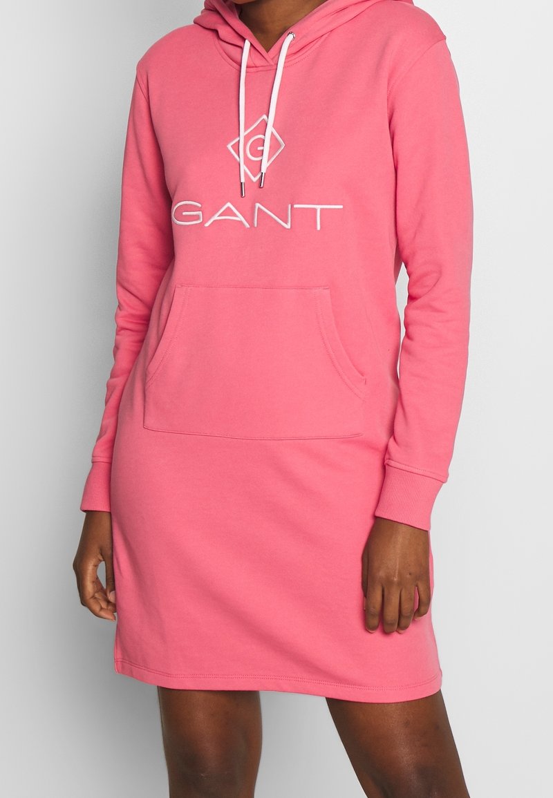 Femme portant une robe à capuche rose à manches longues avec poche avant et logo blanc "GANT" sur la poitrine.