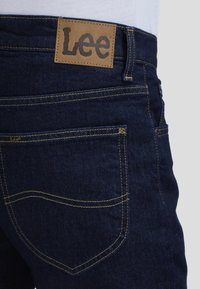 Primo piano della tasca posteriore di jeans denim blu scuro con cuciture gialle e toppa in pelle beige del marchio Lee in vita.